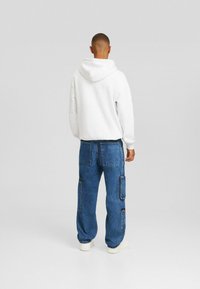 Sudadera blanca con capucha combinada con pantalones cargo de mezclilla azul que cuentan con múltiples bolsillos y costuras en contraste. Modelo de espaldas a la cámara.