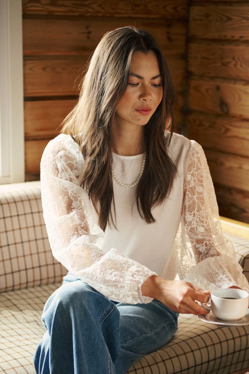 Jeune femme aux longs cheveux portant un haut blanc à manches en dentelle et un jean, assise sur un canapé à carreaux, tenant une tasse de café.