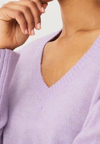 Maglione viola chiaro con scollo a V, polsini e scollo a coste, realizzato in materiale morbido e dalla texture soffice. Primo piano dello scollo e della zona della spalla.