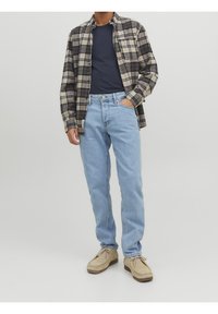 Man i ljusblå jeans, beige snörskor, marinblå t-shirt och en ofodrad beige och svart rutmönstrad skjorta, stående med en hand i fickan.