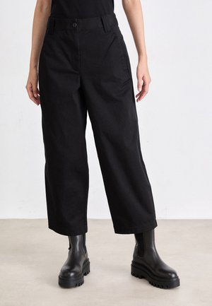 Pantaloni neri a gamba larga con chiusura frontale a pulsante, tasche laterali e una texture liscia, abbinati a stivaletti neri con suola spessa.