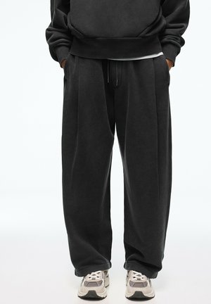 Person iført løstsiddende sorte joggingbukser og sort sweatshirt med hænderne i lommerne, stående med lysefarvede sneakers.