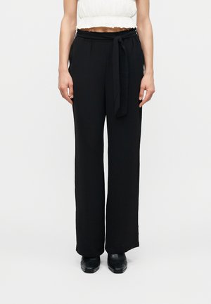 Femeie purtând pantaloni negri largi, cu talie înaltă și curea legată în talie, asortați cu pantofi negri și un top alb scurt.