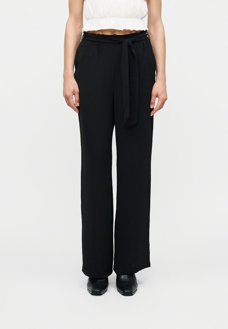 Γυναίκα που φοράει μαύρο παντελόνι με φαρδύ πόδι και ψηλή μέση, με ζώνη δεμένη στη μέση, συνοδευόμενο από μαύρα παπούτσια και λευκό κοντό τοπ.