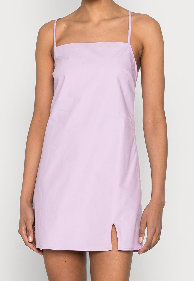 Femme portant une mini robe légère violette sans manches avec de fines bretelles et une petite fente sur le côté avant gauche, debout devant un fond uni.