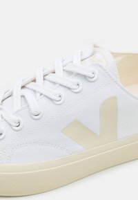Hvit kanvas sneakers med en beige logo, rund gummitå og flate snører. Den teksturerte sålen gir grep og holdbarhet.