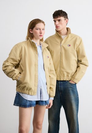 Ung mand og kvinde iført beige jakker med lynlås og elastiske manchetter, en blå denimnederdel og jeans, poserende mod en ensfarvet hvid baggrund.