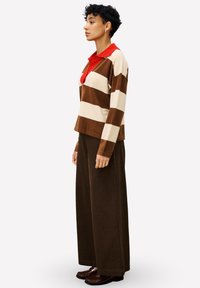 Gestreepte trui in bruin en crème met een rode kraag, gecombineerd met brede bruine broeken. De outfit heeft een ontspannen pasvorm en zachte stof.