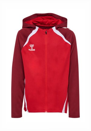 Ung pige stående iført en rød og mørkerød hættetrøje med lynlås, matchende røde shorts, hvide crew-sokker og hvide sneakers på en ensfarvet baggrund.