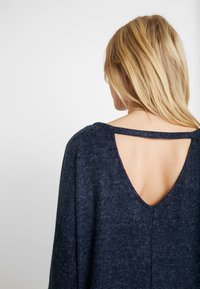 Femme blonde de dos portant un pull bleu foncé avec un large dos ouvert en forme de V et une fine sangle horizontale traversant le haut du dos.