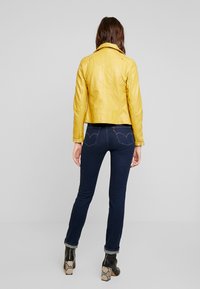 Giubbotto in pelle giallo dal design aderente, abbinato a jeans blu scuro e stivaletti neri alla caviglia con tacco decorato.