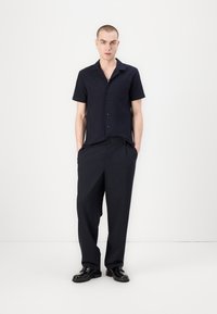 Camicia a maniche corte di colore blu scuro, con bottoni e taschino, abbinata a pantaloni a righe verticali a vita alta e scarpe nere. Texture liscia.