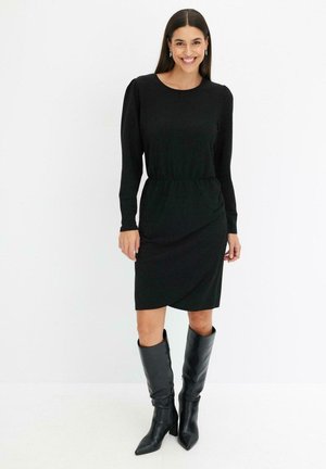 Abito lungo nero a maniche lunghe con vita cinte e dettagli drappeggiati, abbinato a stivali neri a punta e alti fino al ginocchio. Texture liscia in tutta la superficie.