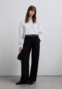 Camisa blanca de manga abullonada, combinada con pantalones negros de talle alto con cinturón plateado con tachuelas. Bolso negro y zapatos planos.