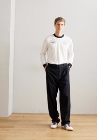 Homme debout à l'intérieur portant une chemise blanche à manches longues avec des logos, un pantalon noir et des baskets blanches avec des rayures noires, les mains dans les poches.