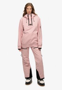 Pink vandtæt jakke med hætte, frontlomme og sorte detaljer, parret med matchende pink bukser og sorte, robuste sneskodder.