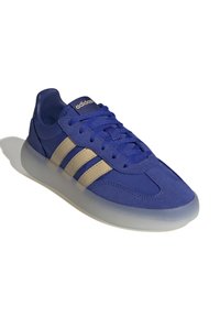 Modré semišové tenisky s hladkou, průhlednou podrážkou. Obsahují tři zlaté pruhy. Zaoblená špička a šněrovací design pro bezpečné uchycení.