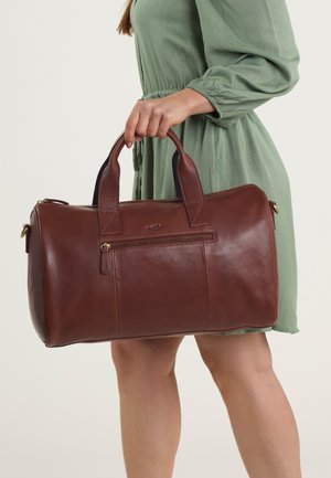 Borsa da viaggio in pelle marrone con due manici rotondi, una chiusura zip sulla parte superiore e una tasca frontale. Presenta una texture liscia e dettagli in metallo dorato.