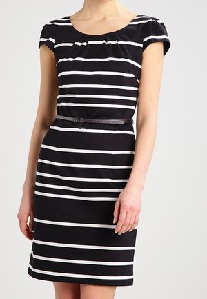 Femme portant une robe noire à rayures horizontales blanches et une fine ceinture noire à la taille, debout devant un fond uni.