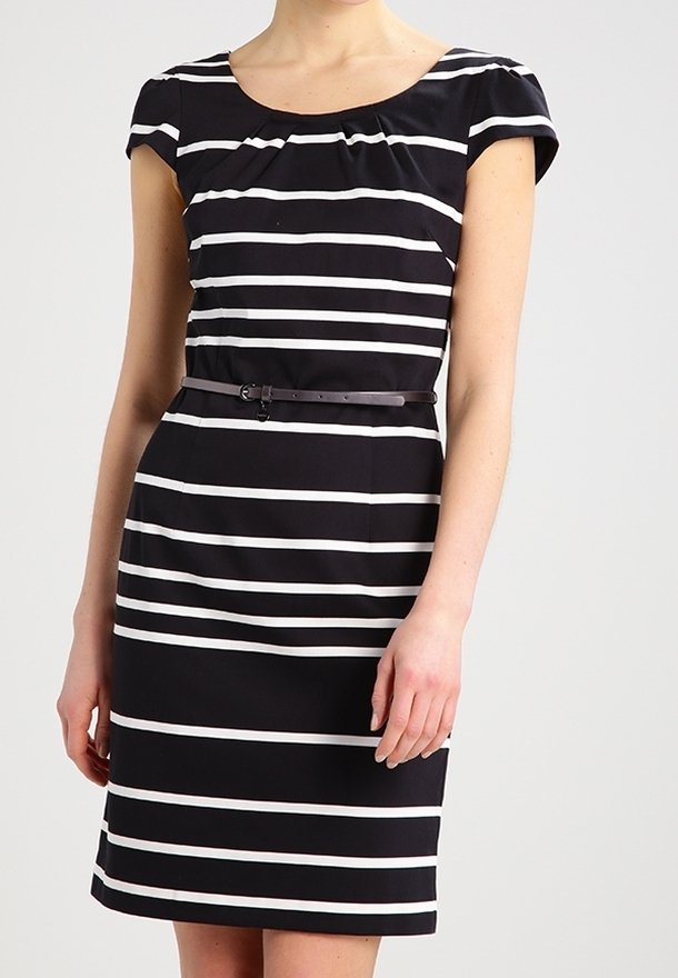 Femme portant une robe noire à rayures horizontales blanches et une fine ceinture noire à la taille, debout devant un fond uni.