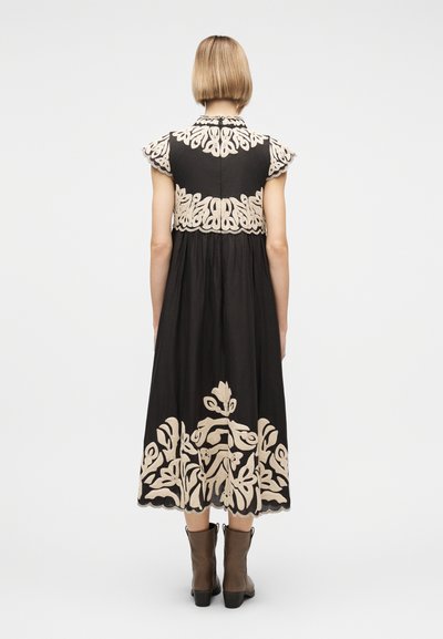 Γυναίκα με κοντά ξανθά μαλλιά φοράει μαύρο μίντι φόρεμα με μπεζ κεντήματα με λουλούδια στην πλάτη και το τελείωμα, συνδυασμένο με καφέ μποτάκια αστραγάλου.