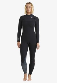 Frau mit blonden Haaren, die einen schwarzen, bodenlangen Neoprenanzug mit einem dunklen gemusterten Einsatz am linken Wadenbereich trägt und barfuß vor einem weißen Hintergrund steht.