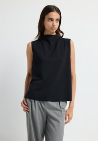Jeune femme aux longs cheveux noirs portant un haut noir sans manches et un pantalon gris plissé sur fond blanc uni.