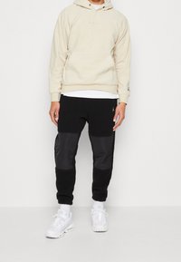 Beige Kapuzenpullover mit Fronttasche und Kordelzug, kombiniert mit schwarzen Hosen aus strukturiertem Stoff und glatten schwarzen Einsätzen, abgerundet mit weißen Sneakers.