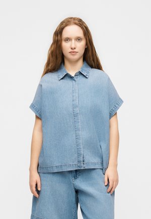 Jeune femme aux longs cheveux ondulés portant une chemise en jean bleu clair ample et un short en jean large assorti, debout devant un fond uni.