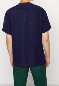 Navy linen short-sleeve shirt with a relaxed fit, back yoke detail, and rounded hem; paired with green pants.