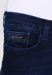 Jeans de mezclilla de color azul oscuro que cuentan con un bolsillo frontal con una etiqueta negra que dice "Calvin Klein Jeans". Textura suave; diseño clásico de cinco bolsillos.