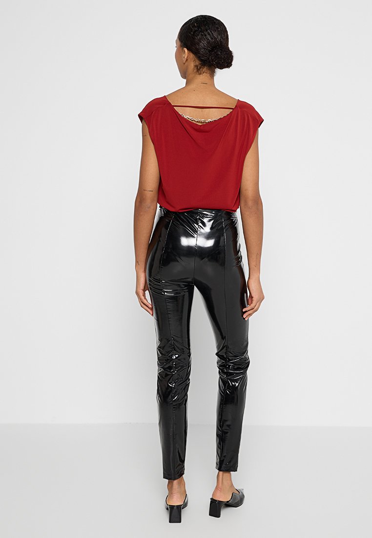 Rotes Top mit tiefem Rückenausschnitt und goldenen Akzenten; schwarze, hochtaillierte, glänzende Hose mit Raffung entlang der Beine; elegantes und figurbetontes Design.