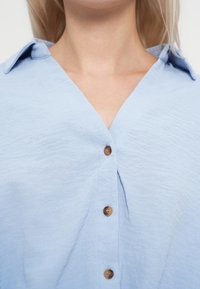 Femme portant une chemise texturée bleu clair à boutons avec un col et trois boutons marron visibles, montrée du cou à mi-poitrine.