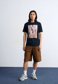 En person står i et rum iført en sort t-shirt med et farverigt grafik, brune cargo shorts og sneakers på et blåt tæppe.