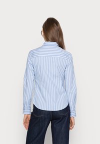 Hellblaues Langarmhemd mit vertikalen weißen Streifen, mit Kragen, knöpfbaren Manschetten und abgerundetem Saum, kombiniert mit dunklen Jeans.