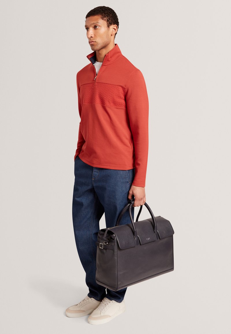 Rotes Sweatshirt mit Reißverschluss und strukturiertem Einsatz, dunkle Jeans aus Denim, cremefarbene Sneaker und eine schwarze Lederhandtasche mit klarer Form und Henkel.