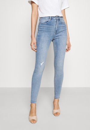 Slim-fit lyseblå jeans med let slid på låret, båret med en hvid top og beige åbne hæle på en ensfarvet baggrund.