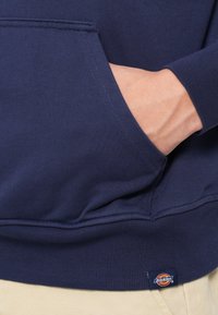 Ναυτικό μπλε φούτερ με ριχτό στρίφωμα και μανίκια. Κεντρική τσέπη με λεπτομέρειες ραφής. Ετικέτα με το λογότυπο Dickies ορατή στο κάτω αριστερό μέρος.