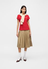 Κόκκινο βαμβακερό μπλουζάκι με λογότυπο, μπεζ πλεκτό κασκόλ δεμένο στο λαιμό, χακί πλισέ φούστα, λευκές κάλτσες και μπορντό ψηλοτάκουνα με μυτερή μύτη.