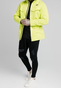 Lysegul, letvægtsjakke med to forlommer, parret med sorte slidte skinny jeans og hvide sneakers.