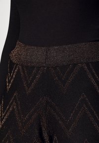 Czarny dzianinowy element garderoby z metalicznymi, brązowymi wzorami w zygzak oraz prążkowanym pasem, noszony z czarnym prążkowanym topem.