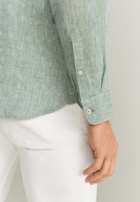 Camicia di lino verde chiaro con una finitura testurizzata, polsini a bottone e orlo arrotondato, abbinata a pantaloni bianchi. Dettagli di cuciture visibili.