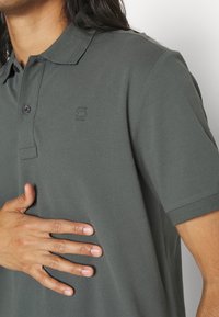 Personne portant un polo gris foncé à manches courtes et posant la main gauche sur la poitrine, montrant les boutons et les détails du col.
