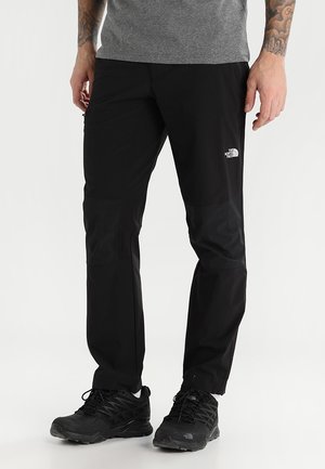 Mann trägt schwarze North Face-Hose und schwarze Wanderschuhe und steht vor einem schlichten weißen Hintergrund.