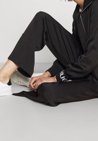 Pantaloni neri a gamba larga realizzati in tessuto morbido, abbinati a una giacca con zip. Sneakers bianche con dettagli neri e logo sui polsini.