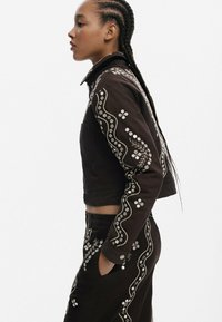 Brązowa krótka kurtka z misternym białym haftem kwiatowym i falistymi wzorami wzdłuż rękawów, w zestawie z pasującymi spodniami.