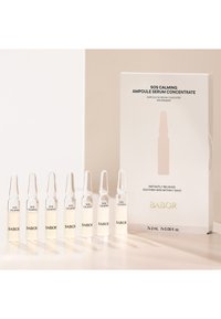 Sju glasampuller fyllda med ljusgult serum, i en låda märkt "SOS Calming Ampoule Serum Concentrate" med svart och guld text.