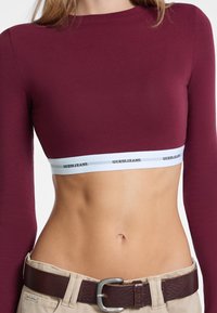 Bordeaux lange mouwen crop top met een witte elastische band met het "GUESS JEANS" logo in het zwart. Gladde textuur, aansluitend ontwerp.