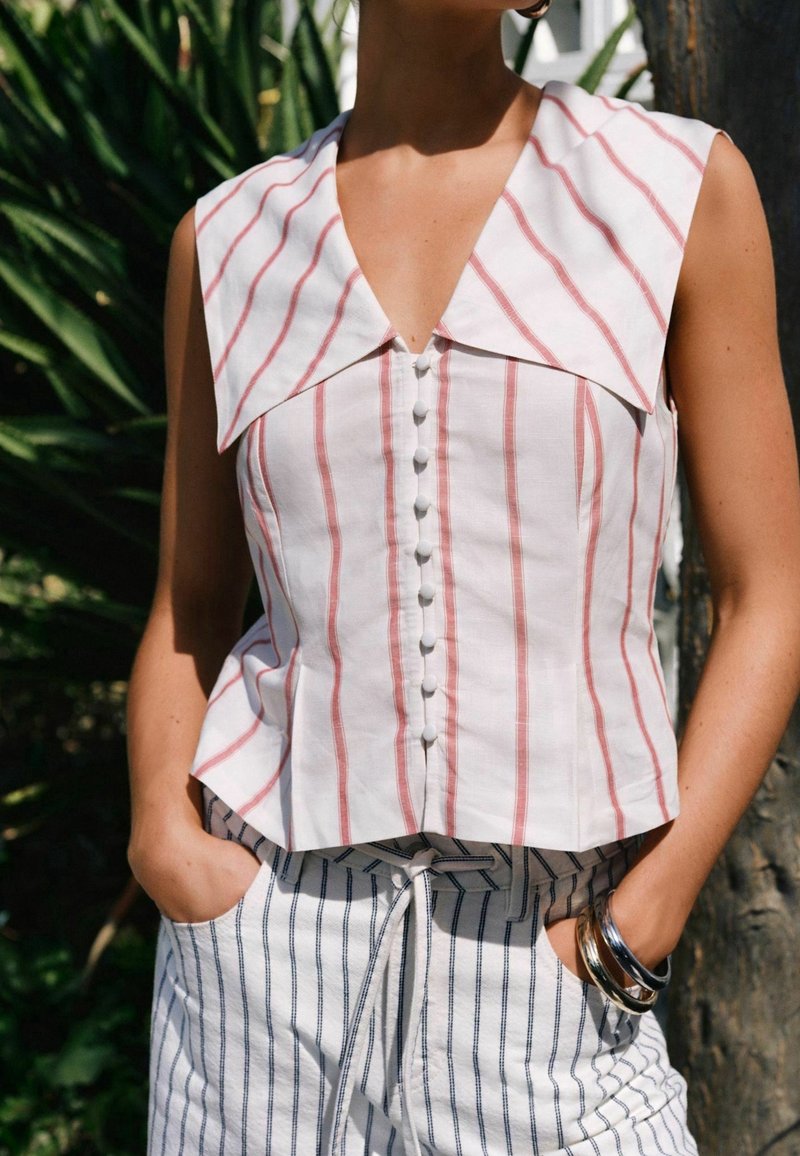 Blouse blanche sans manches avec des rayures verticales rouges et un grand col, associée à un pantalon blanc taille haute avec des rayures noires et un lien à nouer devant.