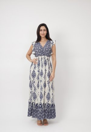 Mujer de pie con la mano en la cadera, vestida con un maxi vestido sin mangas blanco y azul marino con estampado, y sandalias, sobre un fondo blanco liso.